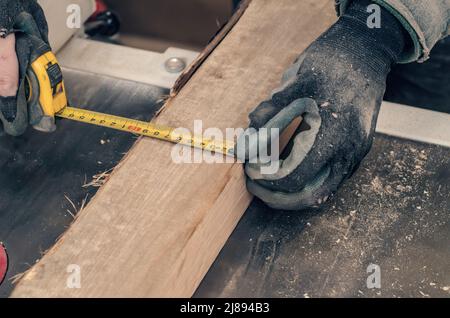 Der Zimmermann misst die Breite der Holzplatte mit einem Maßband. Hände des Zimmermanns in Handschuhen. Stockfoto