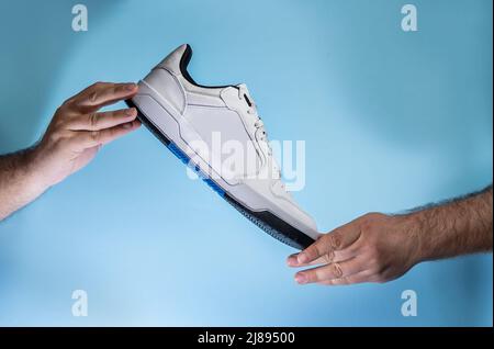 Hände mit neuen Tennisschuhen ohne Marke auf blauem Hintergrund. Neue Turnschuhe oder Sportschuhe ohne Marke – Herren Sportschuhe. Paar Sportschuhe. Stockfoto
