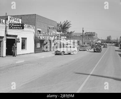 Hauptstraße von Nyssa, Oregon. Samstagnachmittag. [Schilder: 'Towne's Garage; Laird's Body Works - Auto Glass; Theater [zeigt Never Say die mit Martha Raye und Bob Hope und kämpft gegen Gringo mit George O'Brien); Drugs Fountain; Shell; Good Year; Wilson Bros Groceries - Dry Goods; Bob's Tavern'. Güterzug am Bahnübergang notieren]. Stockfoto