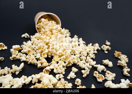 Popcorn wird aus einem braunen Papierbecher auf schwarzem Hintergrund in Nahaufnahme ausgestreut Stockfoto