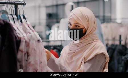 Junge arabische Frau Kunde Käufer in Hijab trägt schützende medizinische Maske im Bekleidungsgeschäft sorgfältig wählt Kleidung auf der Suche nach geeigneten Outfit suchen Stockfoto