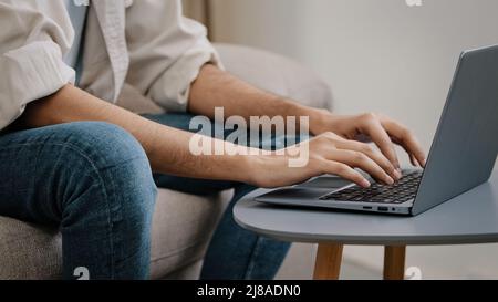 Nahaufnahme der männlichen Hände, die auf der Laptop-Tastatur tippen. Unkenntlich unbekannter Geschäftsmann in Jeans Casual Shirt sitzt auf dem Sofa mit Computer arbeiten Stockfoto