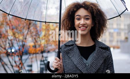 Lächelnd fröhlich afroamerikanisch 20s Modell Tourist girl Ausländer Frau mit transparentem Regenschirm in der Stadt stehend genießen regen reisen in neu Stockfoto