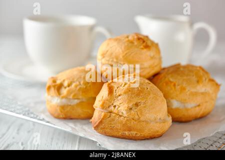 Köstliche hausgemachte Profiterolen oder Ecolirs auf einem Metallgitter, das mit weißem Papier bedeckt ist. Im Hintergrund befindet sich eine Teetasse und ein Milchkännchen. Nahaufnahme. Stockfoto