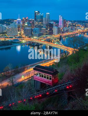 Ein rot-violettes Auto klettert nachts auf die historische Duquesne Incline Railway mit dem Monongahela River und der Skyline von Pittsburgh im Hintergrund Stockfoto