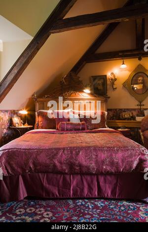 Queen-Size-Bett mit violettem Samt-Bettüberwurf im Hauptschlafzimmer im oberen Stockwerk im alten rekonstruierten Blockhaus im Stil von 1800s Canadian Cottage. Stockfoto