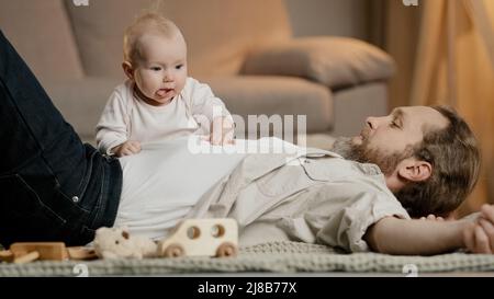 Kaukasischer Familienvater mit kleiner Tochter Sohn liegen auf dem Boden zu Hause spielen. Baby girl junge neugeborenes Kind kriecht über Vater Kinderbetreuung Vaterschaft Stockfoto