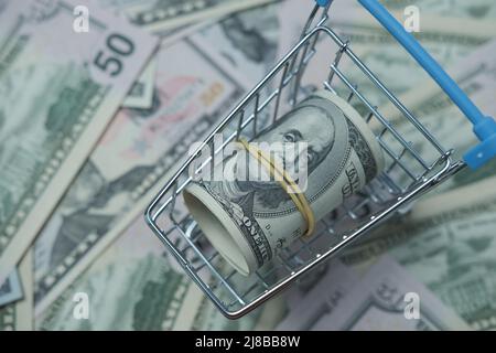 In einen Schlauch gerollte Dollars liegen in einem Einkaufskorb. Der Korb steht auf einem verschwommenen Geldhintergrund. Stockfoto