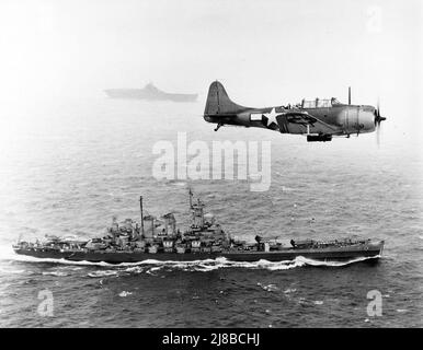 U.S. Navy Douglas SBD-5 die untadelige Bombenschwadron VB-16 fliegt auf dem Weg zur Invasion der Gilbert-Inseln, 12. November 1943, eine unterseeboot-Antipatrouille tief über dem Schlachtschiff USS Washington (BB-56). Das Schiff im Hintergrund ist die USS Lexington (CV-16), der Heimatträger des Flugzeugs. Stockfoto