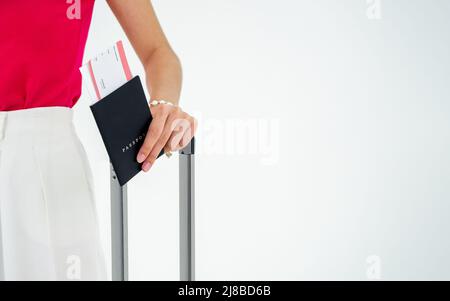 Nahaufpassung mit Flugticket in der Frauenhand in rosa ärmelloser und weißer Hose, während sie einen auf Weiß isolierten Koffergriff in der Hand hält Stockfoto
