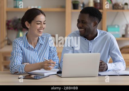 Fröhliche, fröhliche zwei verschiedene Mitarbeiter, die am Arbeitsplatz ihren Laptop teilen Stockfoto