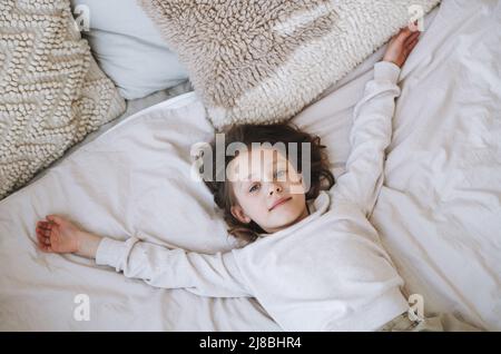 Nettes lustiges kleines Mädchen mit langen Haaren in grauem Heimkleid mit Spaß liegt auf dem Bett zu Hause, Draufsicht Stockfoto