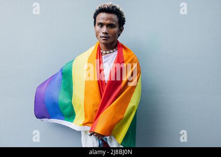 Junger Mann, eingewickelt in eine LGBT-Flagge, der draußen an einer grauen Wand steht Stockfoto