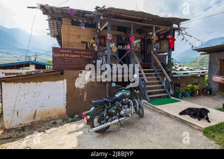 Paro, Bhutan - 24. Oktober 2021: Kunstgeschäft in einem alten, eher heruntergekommenen traditionellen Haus. Das indische Royal Enfield Motorrad und ein schwarzer Hund stehen vorne Stockfoto