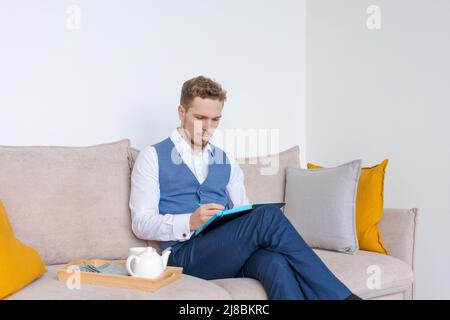 Nachdenklicher Geschäftsmann sitzt mit Tagebuch in der Hand auf dem Sofa, denkt in einem blaubärtigen Anzug über Pläne nach. Plant seine täglichen Routine- und Geschäftstreffen. Konzept für die Aufbewahrung und Planung von Aufzeichnungen Stockfoto
