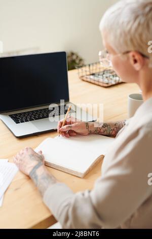 Überblicken Sie die Geschäftsfrau, die am Tisch sitzt und Informationen aufschreibt, während Sie sich den Marketingkurs auf einem Laptop ansehen Stockfoto