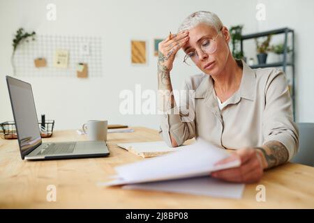 Verwirrt Geschäftsfrau mittleren Alters in runden Brillen reiben Stirn, während Papierkram im Büro Stockfoto