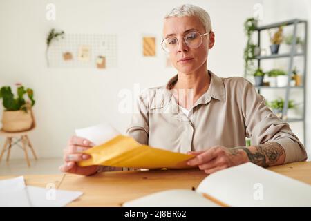 Seriöse blonde Geschäftsfrau mit Brille sitzt am Schreibtisch und öffnet den Umschlag mit einem Geschäftsbrief Stockfoto