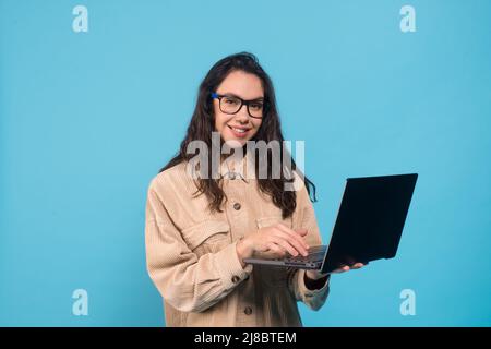 Lächelnde tausendjährige europäische Frau Brünette Student in Brille tippen auf Laptop isoliert auf blauem Hintergrund Stockfoto