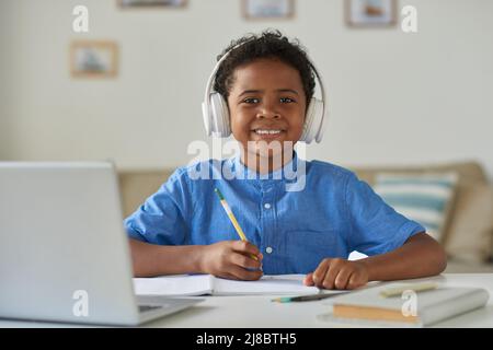 Porträt eines lächelnden schwarzen Jungen in blauem Hemd, der am Schreibtisch sitzt und Notizen im Arbeitsbuch macht Stockfoto