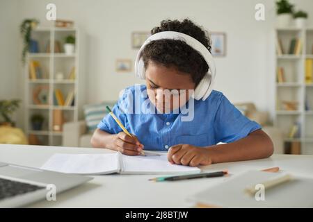 Ernsthafter afroamerikanischer Schuljunge mit Kopfhörern, der am Schreibtisch sitzt und sich Notizen macht, während er online dem Lehrer zuhört Stockfoto