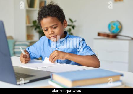Neugieriger schwarzer Schuljunge mit lockigem Haar, der am Tisch sitzt und Notizen im Arbeitsbuch prüft, während er Hausaufgaben macht Stockfoto