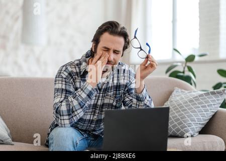 Trauriger, müder, tausendjähriger Kaukasianer mit Stoppeln sitzt auf dem Sofa, der Laptop zieht die Brille ab und leidet unter Kopfschmerzen Stockfoto