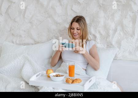 Fröhliche hübsche junge blonde europäische Frau macht Foto von nützlichem Frühstück im Bett allein im weißen Schlafzimmer Stockfoto