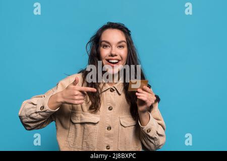Glücklich überrascht lustige junge kaukasische Brünette Dame mit offenem Mund zeigt Finger auf Kreditkarte Stockfoto