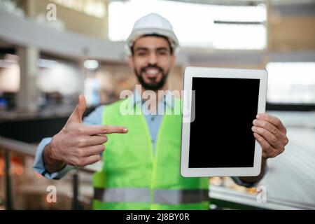 Glücklich attraktiven jungen arabischen Kerl Ingenieur mit Bart in Hut und schützende Uniform zeigt Finger auf Tablette Stockfoto