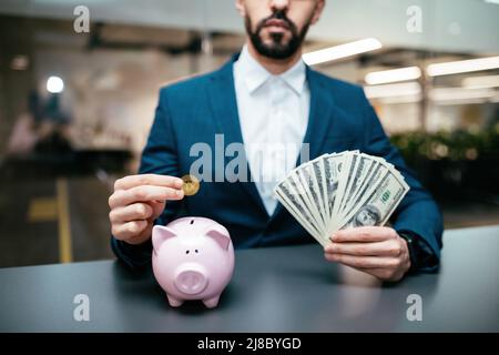 Serious smart Millennial arab guy mit Bart im Anzug hält eine Menge Dollar und setzt Bitcoin in Sparschwein im Büro Stockfoto