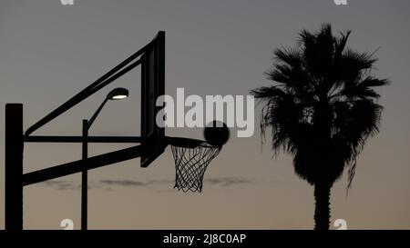 Reifen, Netz und Backboard für Basketball-Spiel Silhouette, Sonnenuntergang Himmel. Basketballplatz auf der Straße, am Strand, Sportplatz oder Spielplatz im Freien, kalifornische Küste, USA. Palme. Nahtloser Cinemagraph mit Schleife. Stockfoto