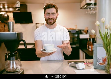 Ein hübscher kaukasischer Barista, der eine Tasse Kaffee vor die Kamera reichte und lächelte. Café-Mitarbeiter. Stockfoto