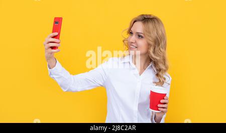 Glückliche blonde Frau mit Kaffeetasse macht Selfie auf dem Telefon auf gelbem Hintergrund Stockfoto