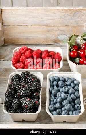 Himbeeren, Blaubeeren, Brombeeren und Kirschen in Kartons auf rustikalem Holztisch. Stockfoto