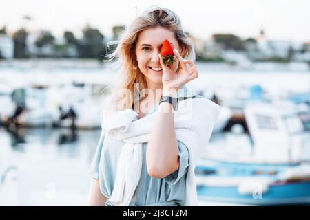 Porträt einer jungen lächelnden Frau, die vor verschwommenem Hintergrund reife, leckere Erdbeeren in der Hand hält und im Resort mit Booten und Yachten entlang des Uferdamms geht Stockfoto
