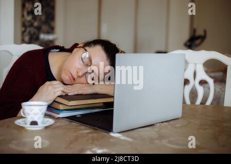 Müde junge Frau in einer Brille, die auf Büchern in der Nähe des Laptops am Schreibtisch im Wohnzimmer schlief. Müdigkeit durch eintöniges Lernen und Ruhe am Arbeitsplatz vom Computer aus Stockfoto