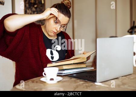 Müde junge Frau, die sich die Brille auszieht, sitzt am Schreibtisch im heimischen Zimmer neben dem Laptop und liest Buch. Müdigkeit durch eintöniges Studium und Computerüberlastung Stockfoto