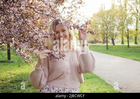 Porträt von lächelnden schönen blonden Haaren Frau in rosa gestrickte Strickjacke hält Zweige blühenden Flieder an sonnigen Tag, Natur Hintergrund. Mode Stockfoto