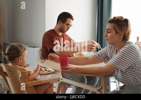 Eltern mit Kindern essen, Frühstück am Tisch. Mutter, die der Familie zu Hause Essen serviert. Essen zusammen mit Kindern. Mädchen im Hochstuhl essen Stockfoto