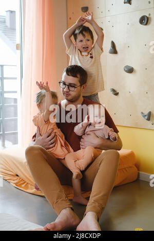Glücklicher Vater mit kleinen Kindern, die zu Hause spielen. Papa kuschelt mit Kindern und hält sie an den Händen. Spaß in der großen Familie, sonniger Tag. DVDAD tragen alle Stockfoto