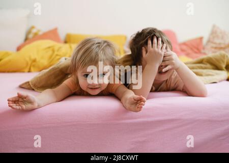 Kinder, die am Morgen auf dem bunten Bett spielen, können sich verstecken. Glückliches Kind liegt auf rosa Bett. Schwester und Bruder im Schlafzimmer, kopieren Raum. Kleinkind Geschwister Stockfoto