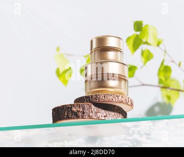 Goldene Kosmetikgläser auf einem Wodden-Podium auf einem Glasregal auf einem natürlichen Frühlingshintergrund mit grünen Ästen. Bio-Kosmetik auf Basis von natürlichen Eingezungenem Stockfoto