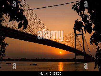 Silhouette von Vidyasagar Setu, Kalkata am Abend. Sonnenuntergang mit dem Boot, das am Fluss Ganges vorbeifährt. Selektiver Fokus. Stockfoto