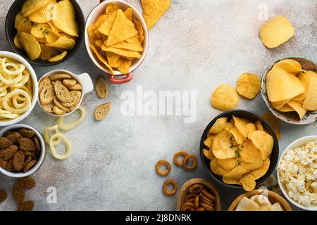 Snacks. Ungesunde Lebensmittel. Alle klassischen Kartoffelsnacks mit Erdnüssen, Popcorn- und Zwiebelringen und gesalzenen Brezeln in Schüsseln auf grauem Beton-Hintergrund Stockfoto