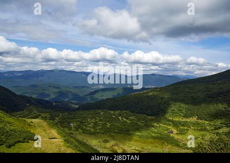 Sonnenbeschienene grüne Senke mit Sträuchern und Bäumen zwischen Berghügeln im Sommer. Oben Blick auf weite Wiese mit Schatten aus den Wolken am blauen Himmel und Bergkette im Hintergrund. Konzept der Landschaft. Stockfoto