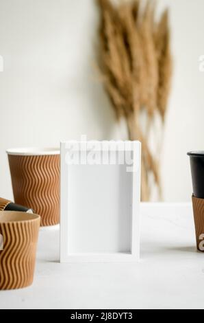 Leerer weißer Rahmen und viele verschiedene Bastelpapier-Einwegbecher. Set von Bastelpappbecher für Kaffee oder Tee auf grauem Hintergrund. Kein Abfall, Kunststoff Fr. Stockfoto