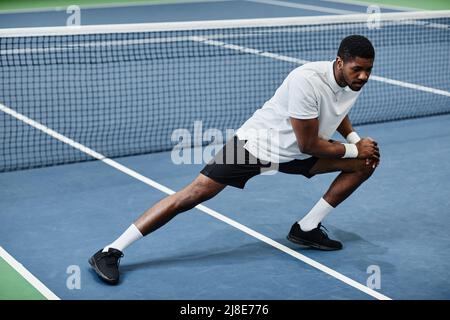 Ganzkörperportrait des jungen schwarzen Sportlers, der sich auf dem Hallentennisplatz ausdehnt und sich aufwärmt Stockfoto