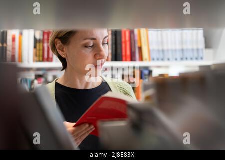 Reife Studentin, die im Bücherregal der Bibliothek nach Büchern sucht, die auf dem Universitätscampus oder College studieren Stockfoto