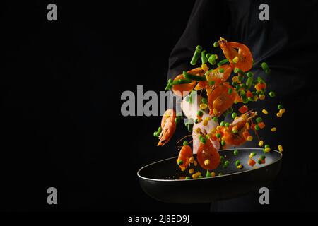 Ein professioneller Koch in schwarzer Uniform bereitet Meeresfrüchte - Garnelen mit Gemüse in einer Bratpfanne zu. Lebensmittel in einem gefrorenen Flug auf schwarzem Hintergrund. Farbe Stockfoto